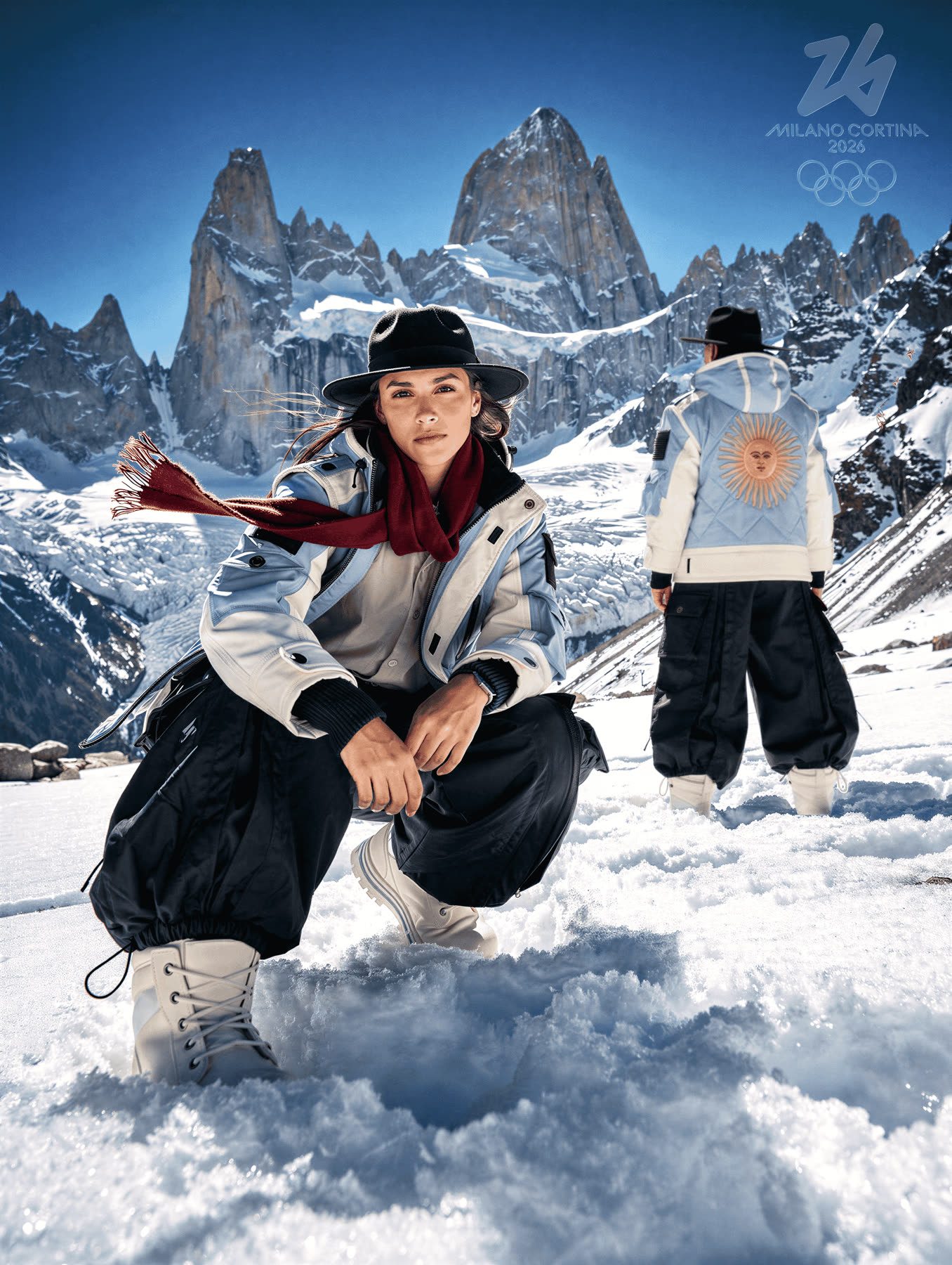 Athlete crouching in snow, mountain range behind, teammate standing further back.