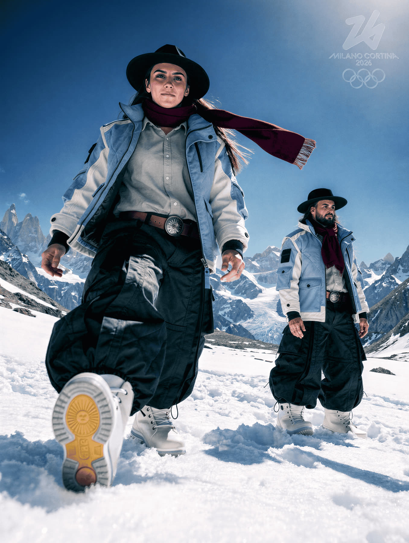 Two athletes in snow, one kneeling with a boot sole toward camera.