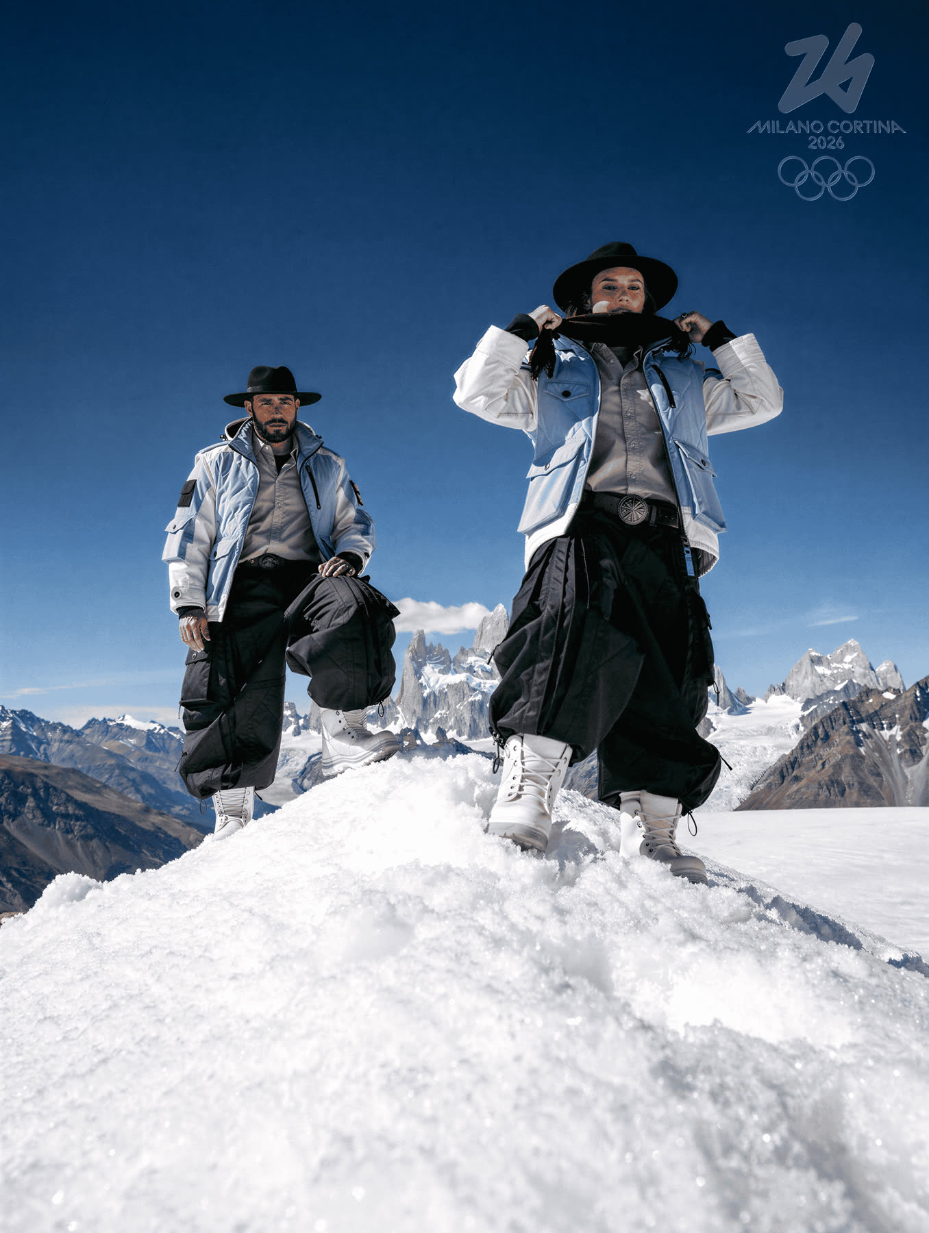 Athlete standing on a snow mound with arms raised, peaks behind.